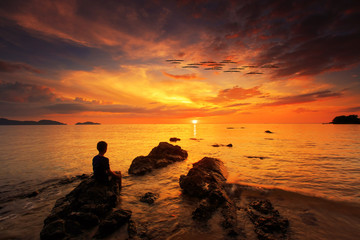 silhouette of young boy show the love hand and standing on rock in the sea with sunset sky, long speed exposure,Tarn-khu beach at Trat, Thailand