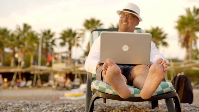 funny middle-aged man is sitting on a sunbed, holding a notebook on lap, smiling and ,wiggling feet