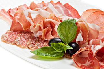 Sliced prosciutto, bacon, salami on a plate decorated leaf of basil and olive.