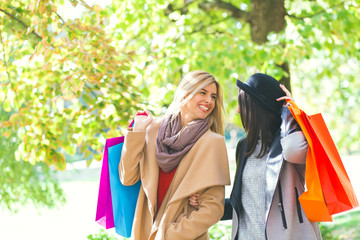 Two smiling young woman with shopping bag in park on sunny autumn day.