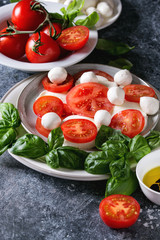 Italian caprese salad with sliced tomatoes, mozzarella cheese, basil, olive oil. Served in ceramic plate with ingredients above over gray texture background. Close up