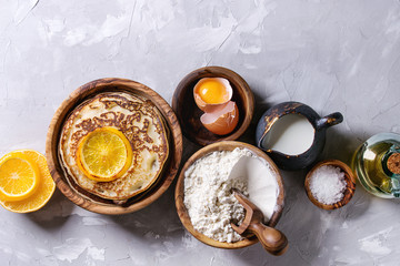Homemade pancakes with fried orange and ingredients above. Wooden bowls of flour, yolk, salt, milk, olive oil over gray texture background. Top view with space. Home cooking concept