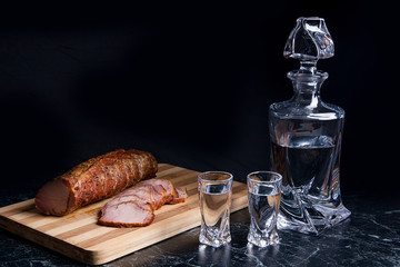 Bottle and shot glasses with vodka with slices of smoked meat on wooden cutting board.