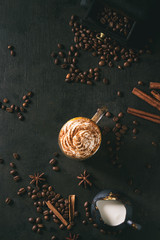 Glass of spicy pumpkin latte with whipped cream and cinnamon standing on black serving board. Coffee beans and spices above. Dark background. Top view, space.