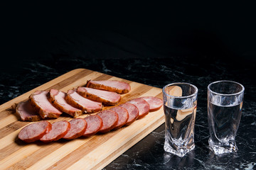 Shot glasses with vodka, slices of smoked meat and smoked sausage on wooden cutting board.