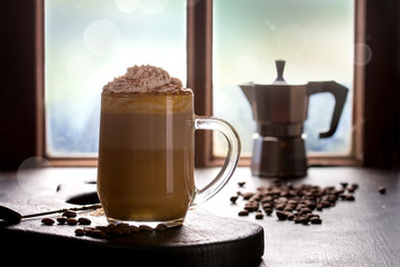 Glass of spicy pumpkin latte with whipped cream and cinnamon standing wooden serving board on cafe table near window with coffeepot and coffee beans above. Atmospheric breakfast