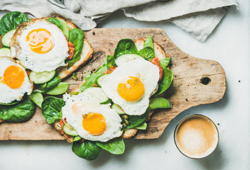 Healthy breakfast sandwiches and cup of coffee. Bread toasts with fried eggs and fresh vegetables on rustic wooden board over grey marble background, top view. Clean eating, healthy, diet food concept