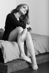 Indoors portrait of attractive young woman in underwear and shirt sitting on bed. Full length. Black and white.
