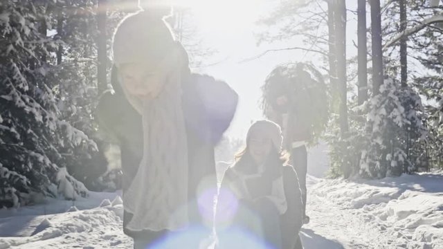 Tracking of cute boy running and pulling laughing mother on sled as bearded father carrying Christmas tree and walking along snowy footpath on sunny day in winter forest