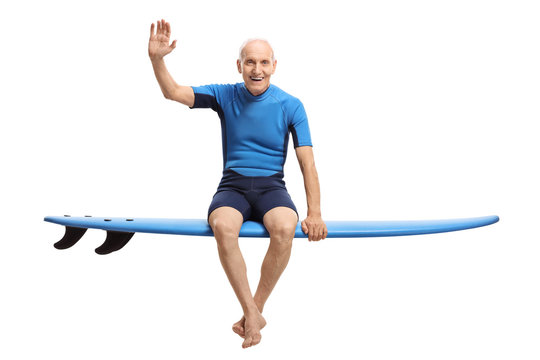 Elderly man in a wetsuit sitting on a surfboard and waving
