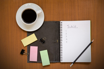 Recycled notebook with To Do List hand writing in the paper note surrounding with black coffee in white cup, yellow, pink & green sticky pads, black pencil & paper clips on dark wood background space
