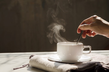 Background of hot black coffee in white ceramic cup saucer over cozy wool scarf stirring for steam by woman right hand over dark background with warm morning light copy space for text or logo insert