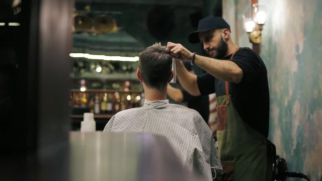 Bearded hairdresser spraying water and combing wet hair of his male client in a retro stylish barbershop. Male hairstyle. Slowmotion shot