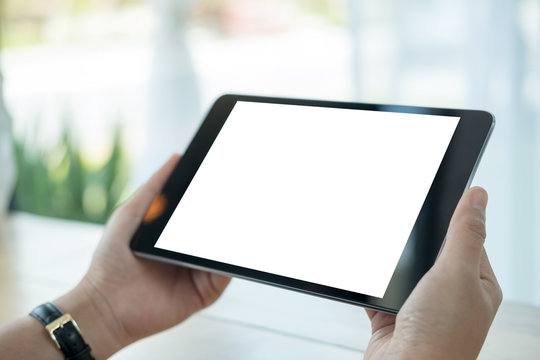 Mockup image of hands holding black tablet pc with blank white screen on wooden table in cafe