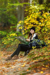 Beautiful brunette tourist girl wears black cap and backpacked have break with map sitting near tree, autumn tourism concept