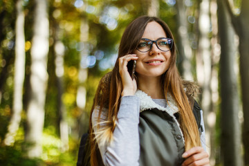 Cheerful brunette tourist backpacked girl have walk through forest holding smartphone, autumn...