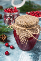 Fresh juicy jam from cranberries in a glass jar on a blue marble background with spruce branches and cones with copy space