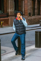 City Life. East Indian American College Student wearing long sleeve T shirt, blue down vest, jeans, leather shoes, sitting on pillar on street in New York, looking up, talking on cell phone, thinking