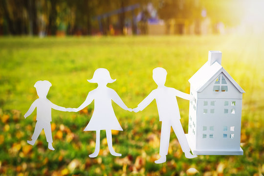 paper family holds hands and house. Family and house concept. On background of green grass on bright sunny day, mom, dad and son from paper hold hands