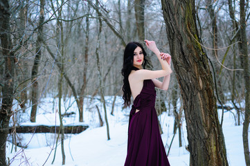 A young woman in an elegant burgundy evening dress in winter among trees and snow conceals a riddle