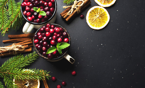 Fresh red cranberries in a white cup, cinnamon sticks, dried orange circles, spruce branches and snowflakes on a black background with copy space top view. Christmas holidays background.