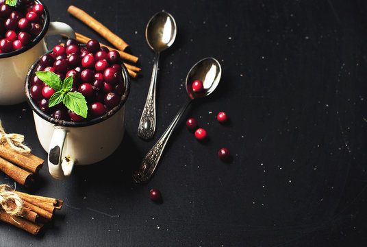 Fresh red cranberries with mint leaves in a white cup, cinnamon and spoons on a dark black background with copy space top view.