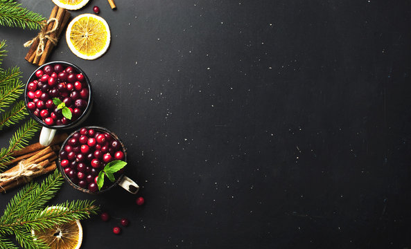Fresh red cranberries in a white cup, cinnamon sticks, dried orange circles, spruce branches and snowflakes on a black background with copy space top view. Christmas holidays background.