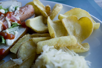 heart shaped fried potato, along with others, on a white ceramic plate of a fast food, in the rest of the dish a hot dog with sauces, wurstel, salad, bread, leaning on a paper tablecloth blue, Italy