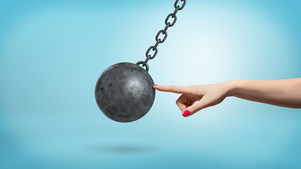 A giant female arm with one finger lightly touching a black iron wrecking ball and stopping its...
