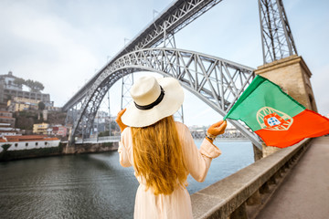 Young woman traveler in sunhat standing back with portuguese flag with the famous iron bridge on...