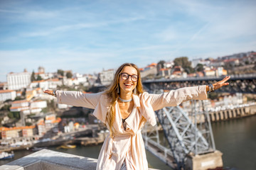 Naklejka premium Portrait of a young woman tourist with raised hands sitting on the beautiful cityscape background with famous bridge in Porto city, Portugal