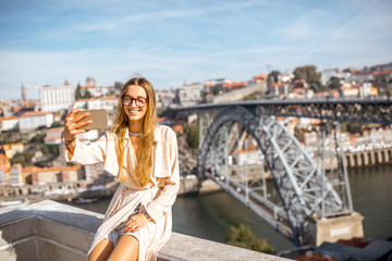 Naklejka premium Young woman making selfie photo with phone on the beautiful cityscape background with famous bridge in Porto city, Portugal