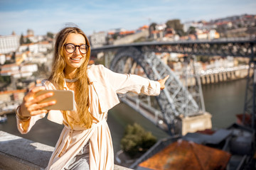 Fototapeta premium Young woman making selfie photo with phone on the beautiful cityscape background with famous bridge in Porto city, Portugal
