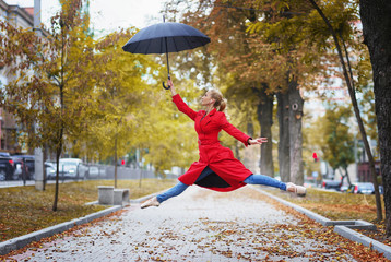 Jumping ballerina with umbrella in one hand. Warm autumn day. Woman in red coat and blue jeans