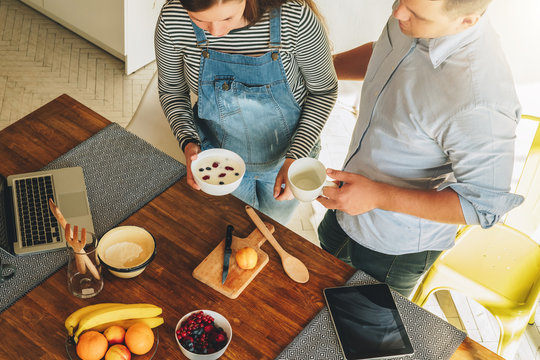 View from above. Young couple in kitchen cooking breakfast. Man is standing near table and drinking tea, his pregnant wife is standing next to him. On table is laptop, tablet computer and fruits.