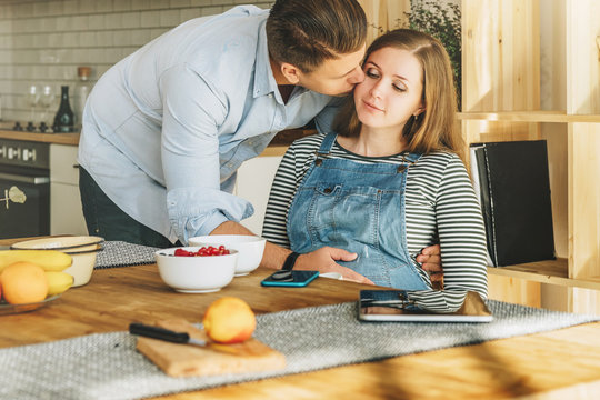 View from above.Morning, breakfast.Young married couple in kitchen.Pregnant woman is sitting at table, man is holding her pregnant belly and kisses.On table is laptop,tablet computer,dishes and fruit.