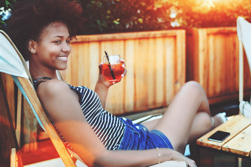 Smiling relaxed young black curly lady with orange glass of delicious alcoholic spritz beverage is...