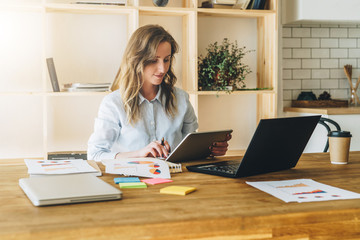 Young businesswoman woman is sitting at kitchen table and uses tablet computer, working, studying. On table laptop, paper documents. Freelancer works at home. Online marketing, education, e-learning.