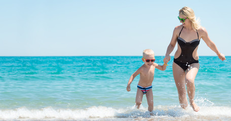 Beautiful young mother and small son holding hands running on the waves on the beach.Fun,family,friendly summer vacation.