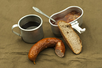a scanty road meal: soup in a mess tin, coffee in a vintage army mug, a loaf of bread and a piece of sausage on a spread tarpaulin
