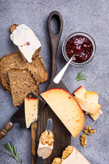 Various types of cheese with wholegrain bread, strawberry jam and walnuts on wooden board on gray stone background. Top view, flat lay.