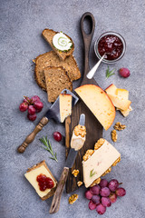 Assorted cheeses on wooden board plate, walnuts, grapes, bread on gray stone background, top view, flat lay.