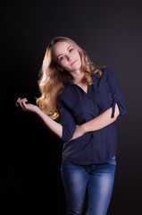 Pretty young blonde model with long wavy hair wears fashionable apparel, posing on a black wall