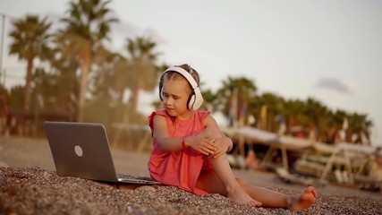 little girl is watching cartoons by laptop and listening sounds by headphones, sitting on a coast - Powered by Adobe