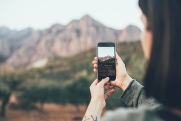 Woman in cosy warm sweater uses smartphone to make photograph of beautiful nature and mountains in park, to share on social media channels to show to friends and family