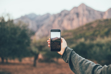 Woman in cosy warm sweater uses smartphone to make photograph of beautiful nature and mountains in park, to share on social media channels to show to friends and family