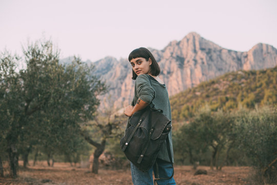 Young female adventurer or tourist explores national park in nature, looks at mountains on sunset, glances at camera from over shoulder, wears comfortable cosy sweater and hipster backpack