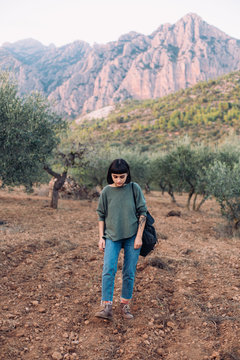 Young woman dressed for an adventure or hike, in blue denim jeans and adventurer hiking boots, walks around vineyard or olive country side field, with mountains in background