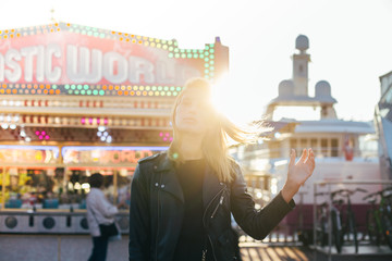 Beautiful fashion model with blonde hair, wears black leather jacket and stands in middle of carnvial, festival or fair, in front of attraction rides during summer time holidays or spring break