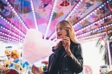 Fashionable young woman in black trendy leather jacket stands in middle of county fair, next to attraction rides or carousel, holds big pink cotton candy or sugar floss, enjoys date night or holidays